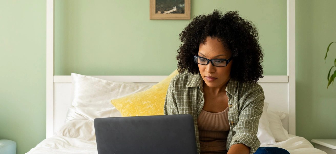 An African American woman works on her laptop while sitting on her bed in a green bedroom.