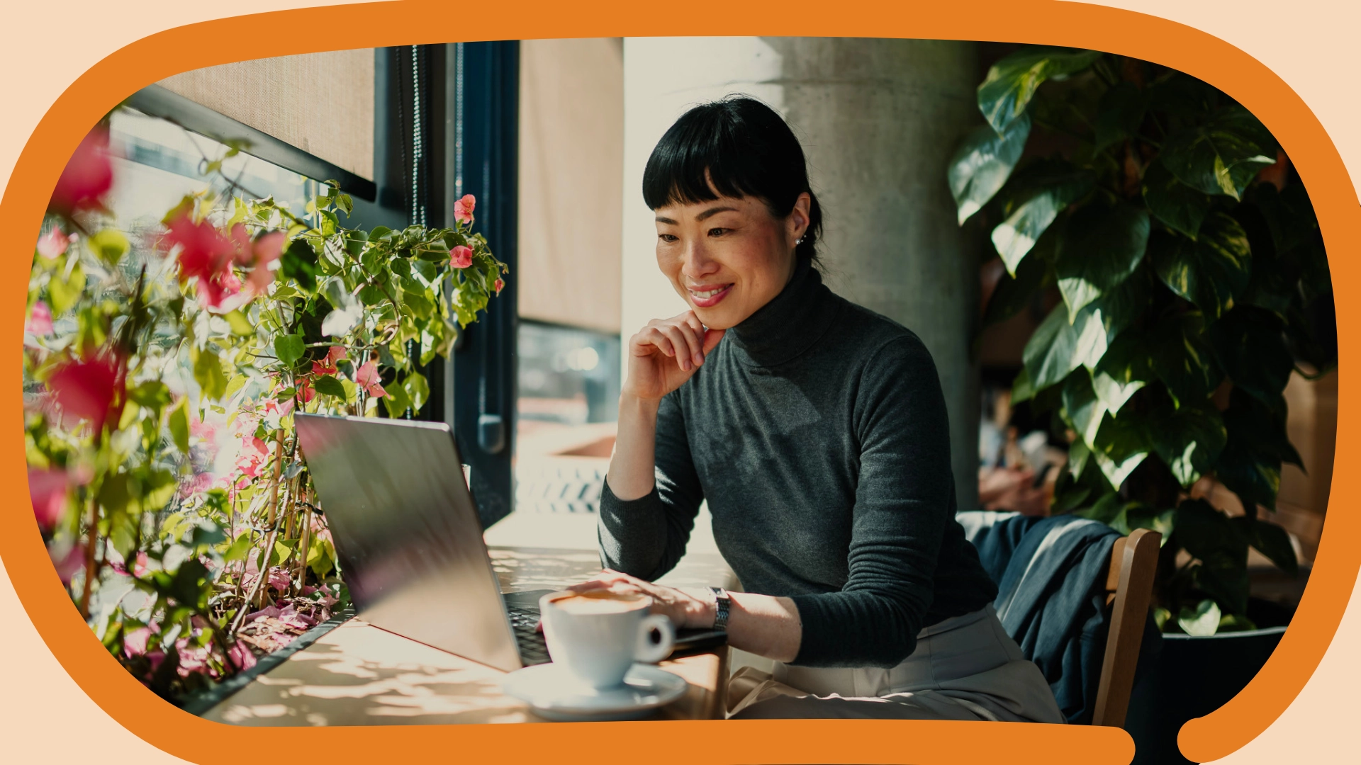 Asian woman smiling while looking at her computer. There is a coffee mug on the desk next to her, and plants lining the room.