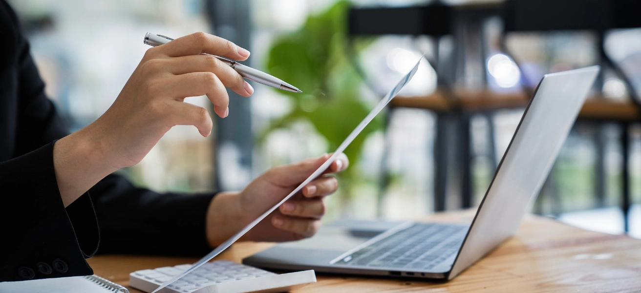 Worker pointing a pen at a piece of paper they hold in their other hand, sitting in front of an open laptop on a light brown desk.