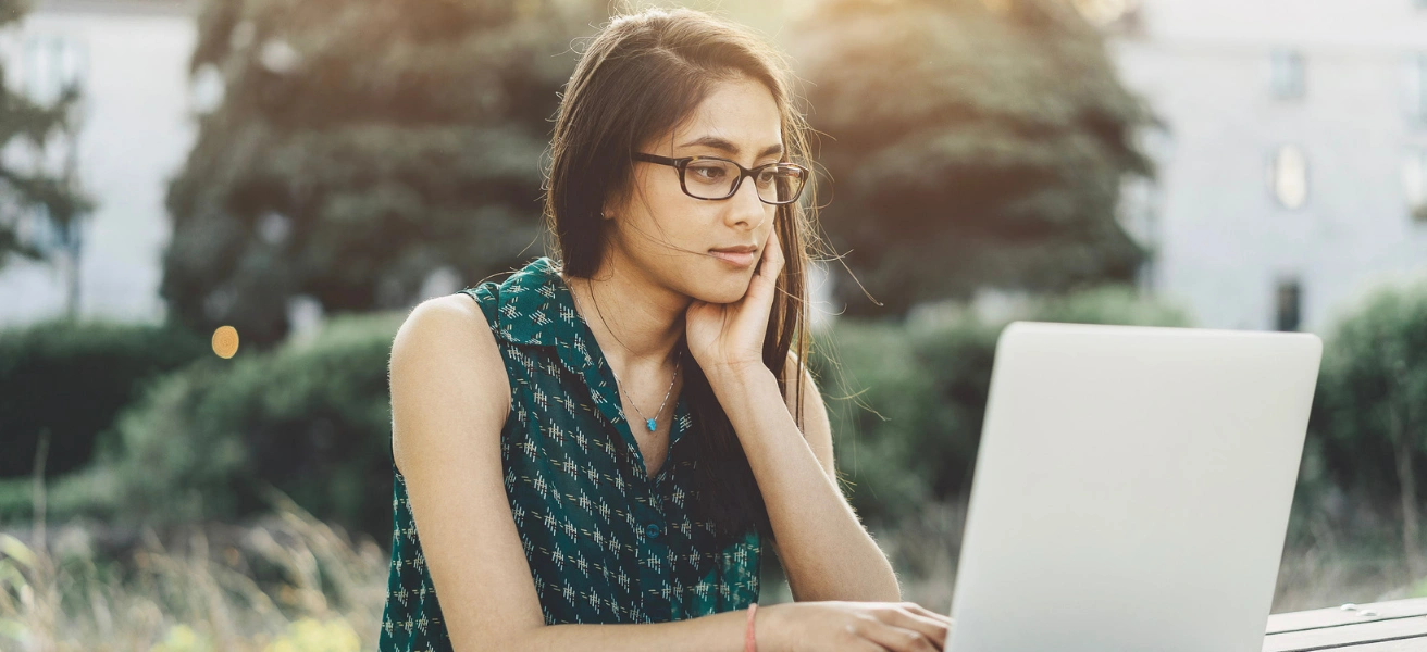 A young woman wearing glasses and a sleeveless top works on her laptop outdoors, looking thoughtful while researching eRank plans for her Etsy shop.